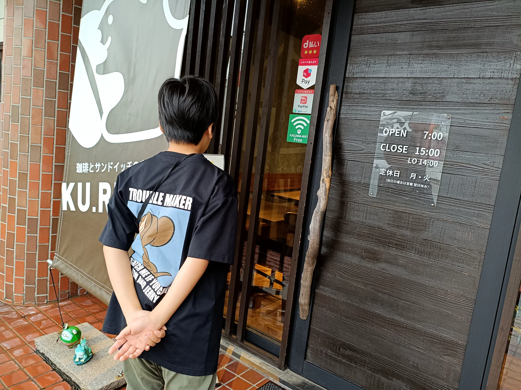 店前で立っている人の後ろ姿と店舗の入口サイン。営業時間のプレートが見える